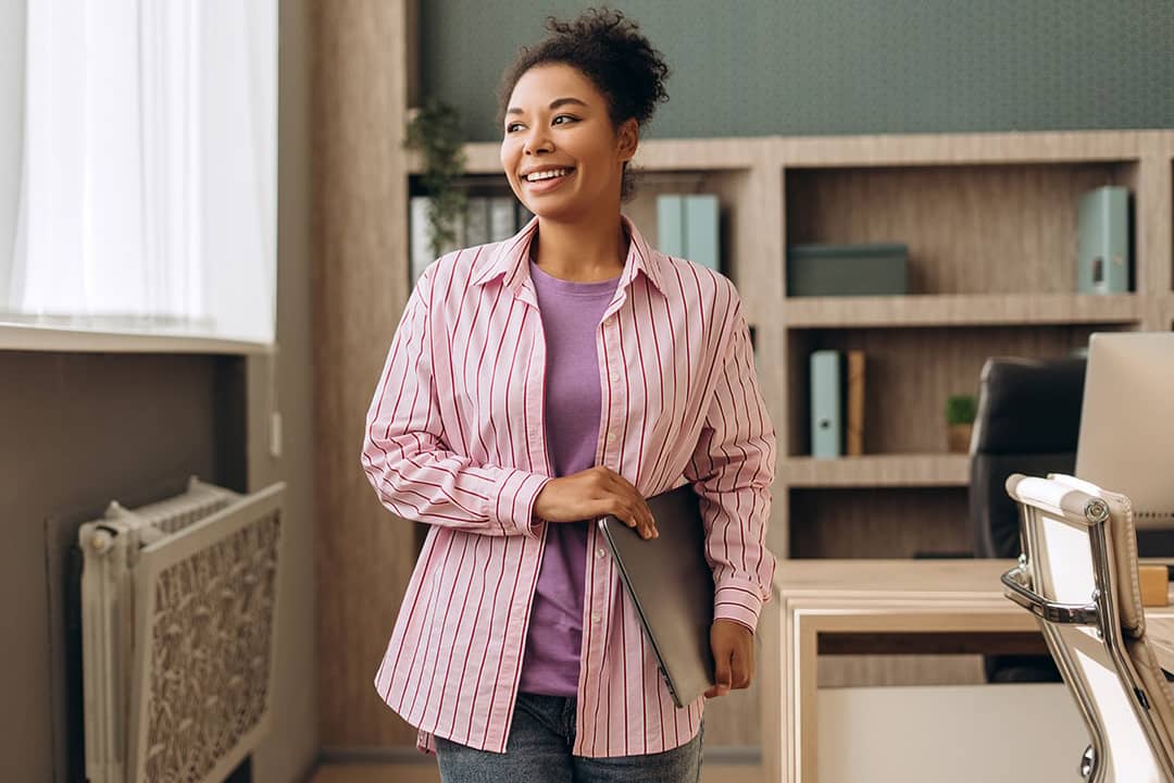 A happy businesswoman in an office walking with her laptop.